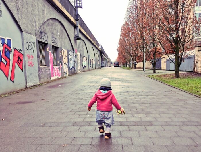 Kleinkind mit Kuscheltier in der Hand läuft langen Weg entlang. Bild von Katharina Mauder