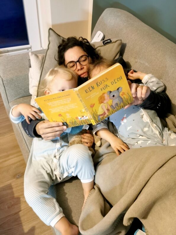 Mama liest ihren Kleinkind-Zwillingen im Liegen auf der Couch ein Pappbilderbuch vor